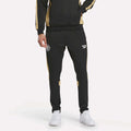 A person showcases only the lower half, wearing Men's MK Dons Travel Pants by Reebok in black and gold with white logos, paired with crisp white sneakers—ideal football fan gear.