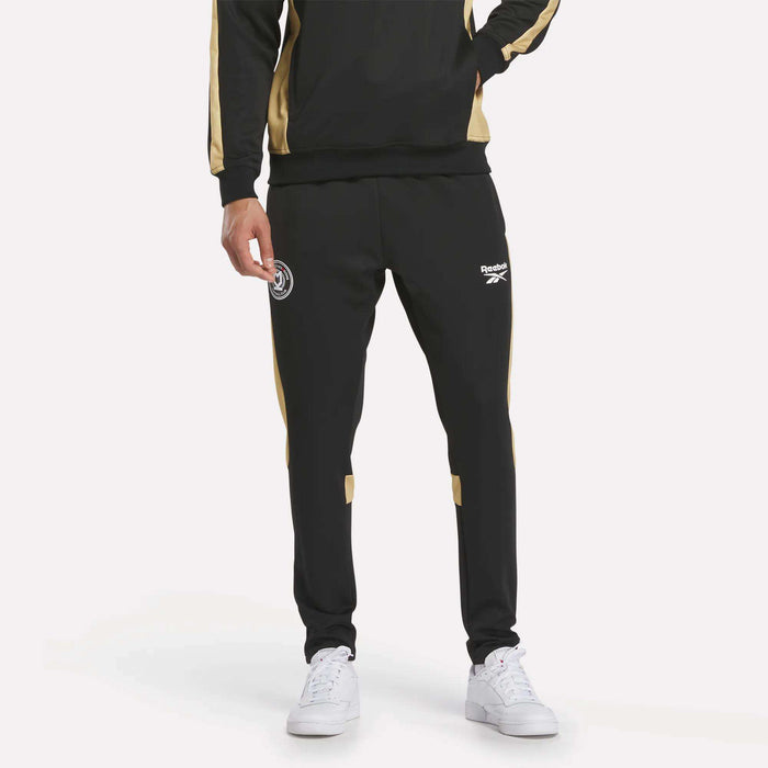 A person showcases only the lower half, wearing Men's MK Dons Travel Pants by Reebok in black and gold with white logos, paired with crisp white sneakers—ideal football fan gear.