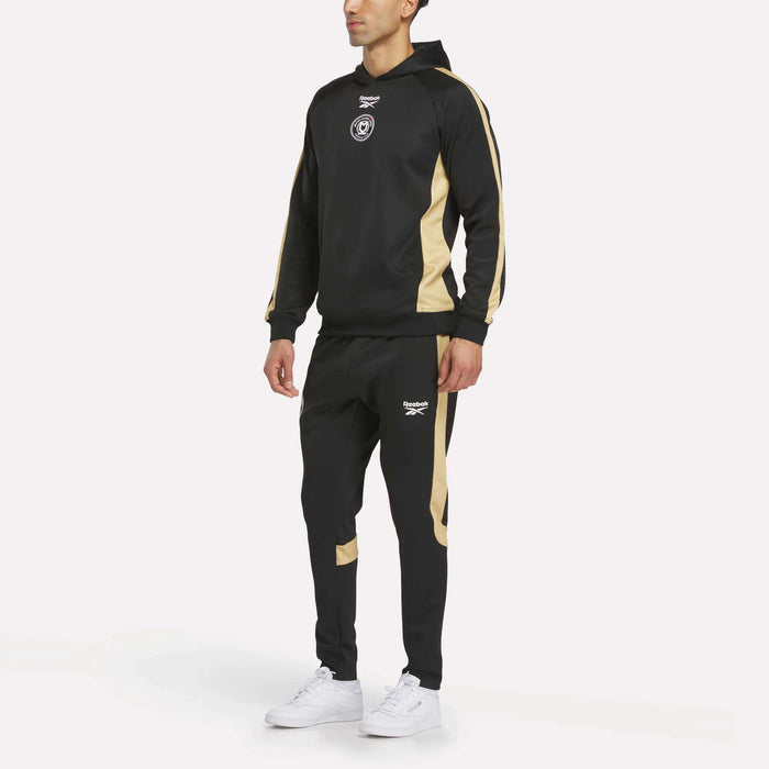 A man wears a black and beige athletic tracksuit with a hoodie and Men's MK Dons Travel Pants by Reebok, both featuring white logos. He completes his look with white sneakers, posed against a plain white background.