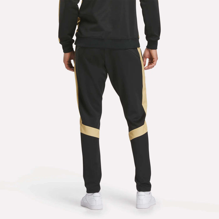 A person is shown from the back against a plain light background wearing Reebok Men's MK Dons Travel Pants with gold side stripes and white sneakers.