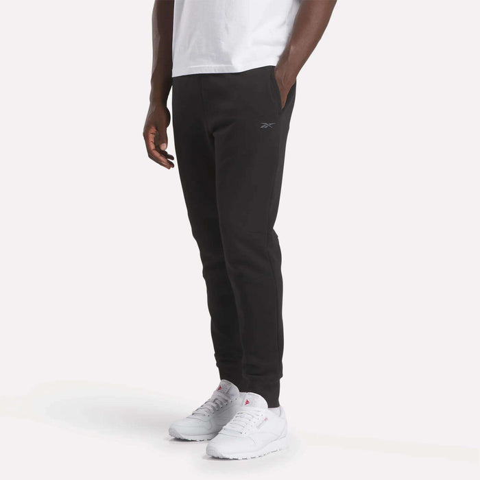 A person wearing a white T-shirt, Reebok Men's Motion Joggers with hands in pockets, and white athletic shoes stands against a plain, light background.