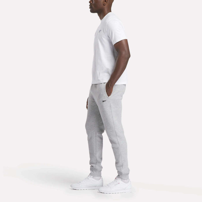 A man stands in profile against a plain white background wearing Reebok Men's Motion Joggers, a white T-shirt, and white athletic shoes.