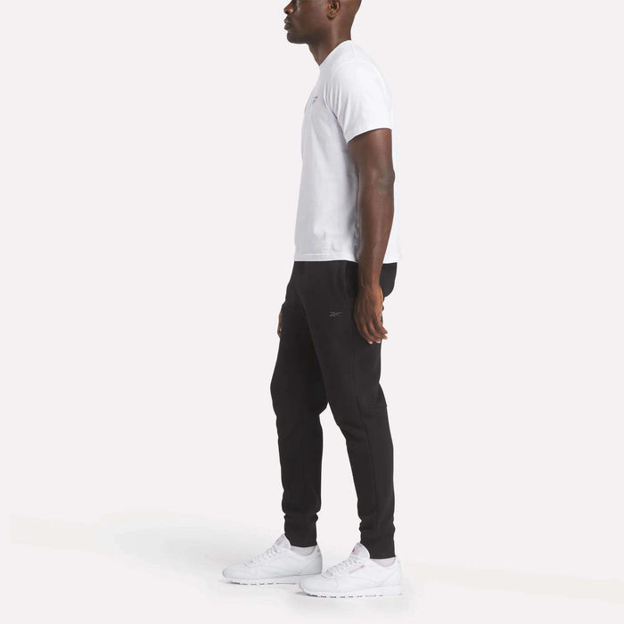 A man stands in profile wearing Reebok Men's Motion Joggers, a white t-shirt, and white sneakers against a plain white background.