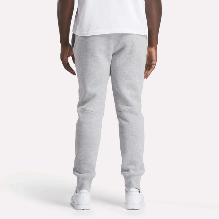 A person stands facing away from the camera, wearing a white T-shirt, Reebok Men's Motion Joggers in light gray slim fit, and white sneakers against a plain light background.
