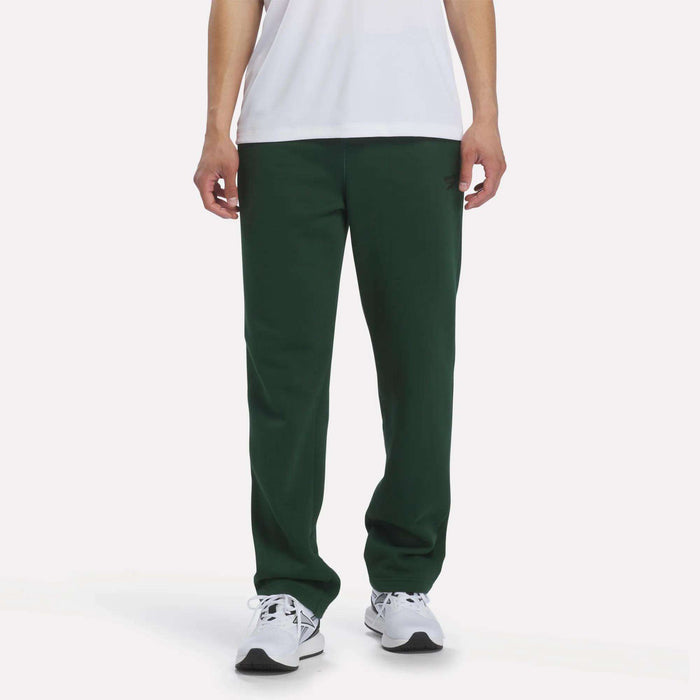 A person stands against a plain white background, wearing a white T-shirt, Men’s Open Hem Pants by Reebok in Heritage Green, and white sneakers. Only the lower half of their body is visible, highlighting the relaxed look of the outfit.