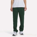 A person stands against a plain white background, wearing a white T-shirt, Men’s Open Hem Pants by Reebok in Heritage Green, and white sneakers. Only the lower half of their body is visible, highlighting the relaxed look of the outfit.