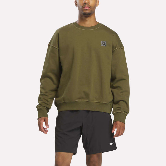 A person wears a relaxed fit Men's Patch Logo Crewneck Sweatshirt by Reebok in Olive Stone, paired with black athletic shorts, standing against a plain white background.
