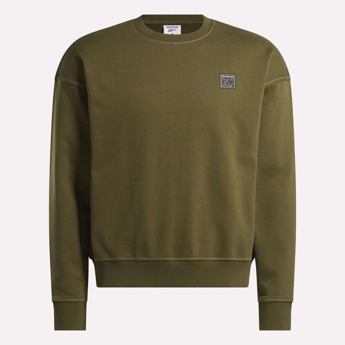 The Reebok Men's Patch Logo Crewneck Sweatshirt features a relaxed fit in olive green with long sleeves, ribbed cuffs and hem, crew neck, and a small square logo patch on the left chest. Displayed against a plain light background.