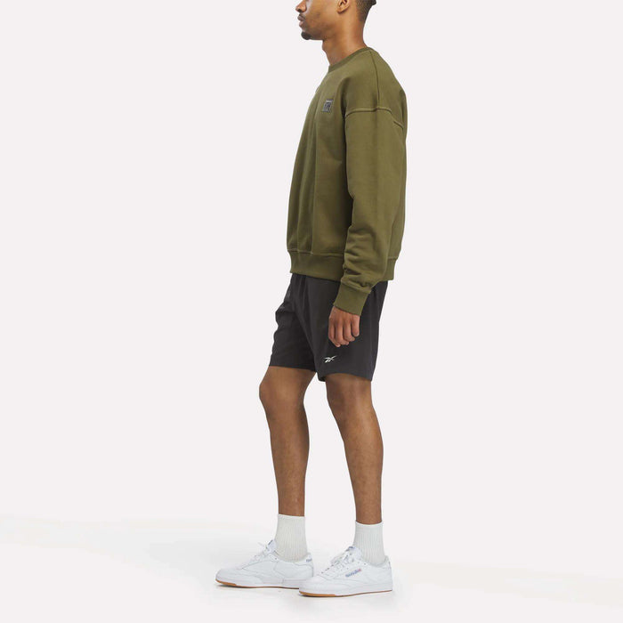A man stands sideways against a plain white background, wearing a relaxed fit Reebok Men's Patch Logo Crewneck Sweatshirt in olive green, paired with black shorts, white socks, and white sneakers.