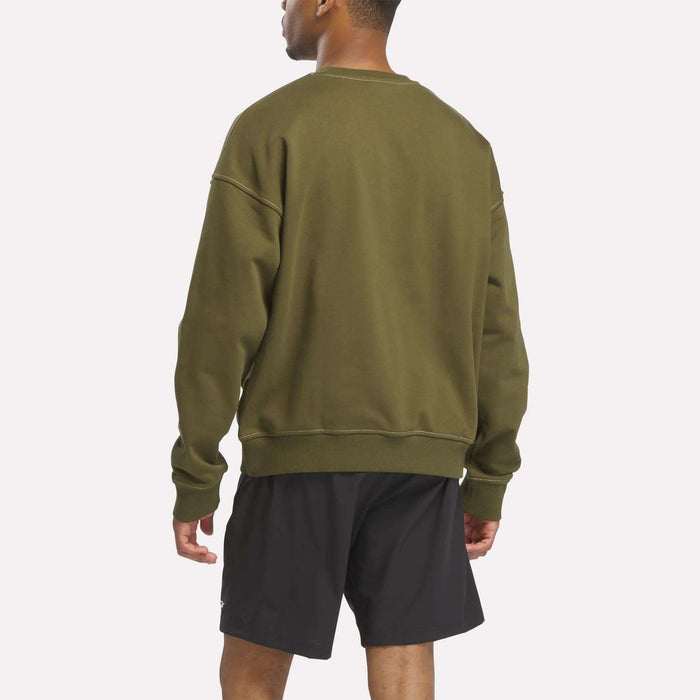 A man faces away, wearing a relaxed fit Reebok Men's Patch Logo Crewneck Sweatshirt in olive green and black shorts, set against a plain white background.