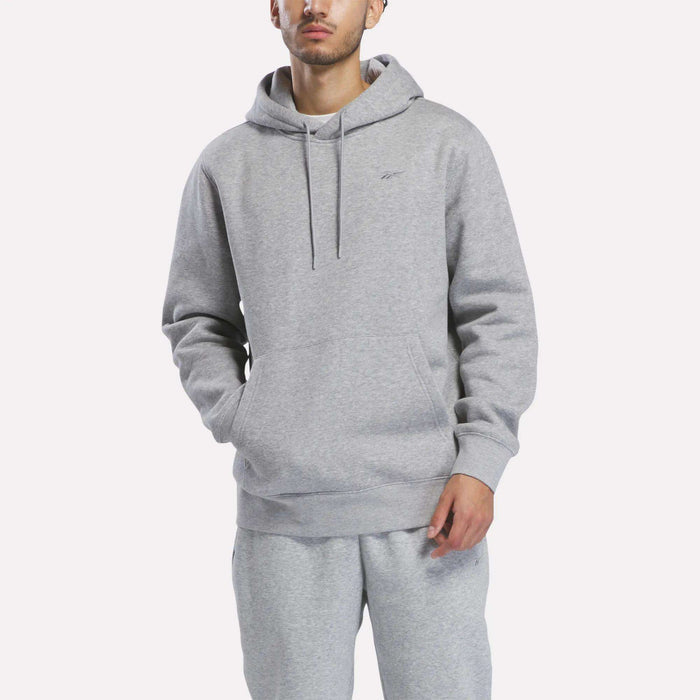 A person wearing the Reebok Men’s Patent Hoodie in Grey Heather, featuring an embroidered logo, and matching sweatpants stands with hands in the hoodie pocket against a plain white background.