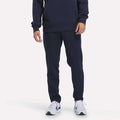 A person is shown from the waist down wearing Reebok Men’s Patent Pant in Vector Navy, featuring a matching navy color and elastic waistband, crafted from soft knit fabric. The outfit is paired with white athletic shoes and photographed against a plain white background.