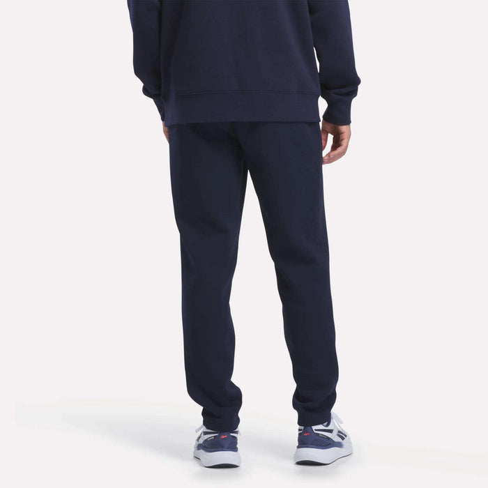 A person stands facing away on a plain light background, showing only the lower half of their body. They are wearing Reebok Men’s Patent Pant and a matching navy blue sweatshirt made from soft knit fabric, featuring an elastic waistband, paired with white and blue athletic shoes.