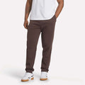 A person is seen from the waist down wearing Reebok Men's Patent Pants in slim fit brown, paired with a white T-shirt and white sneakers with gum soles, standing hands-in-pockets against a plain light background.