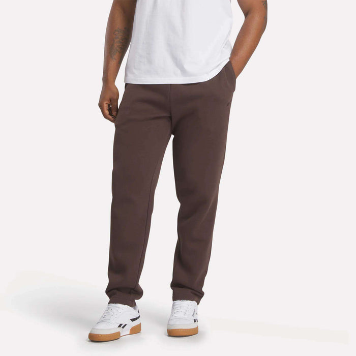 A person is seen from the waist down wearing Reebok Men's Patent Pants in slim fit brown, paired with a white T-shirt and white sneakers with gum soles, standing hands-in-pockets against a plain light background.