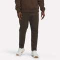 A person is shown from the waist down, wearing Reebok Men’s Patent Pants in Espresso—a slim fit style with an adjustable waistband—paired with a matching sweatshirt and white sneakers, standing against a plain, light background.