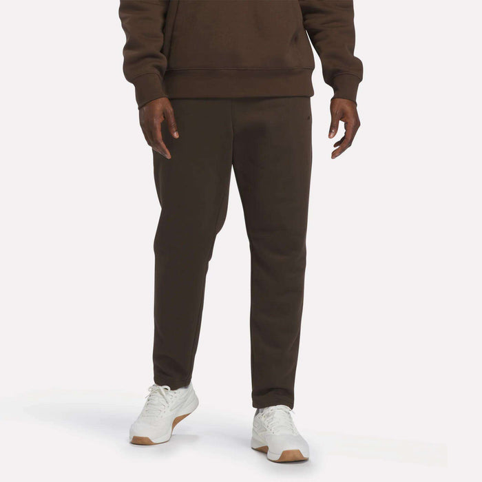 A person is shown from the waist down, wearing Reebok Men’s Patent Pants in Espresso—a slim fit style with an adjustable waistband—paired with a matching sweatshirt and white sneakers, standing against a plain, light background.