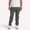 A person wearing a white t-shirt, Reebok Men's Patent Pants in slim fit olive green with an adjustable waistband, and white sneakers stands against a plain light background; their face is not visible.