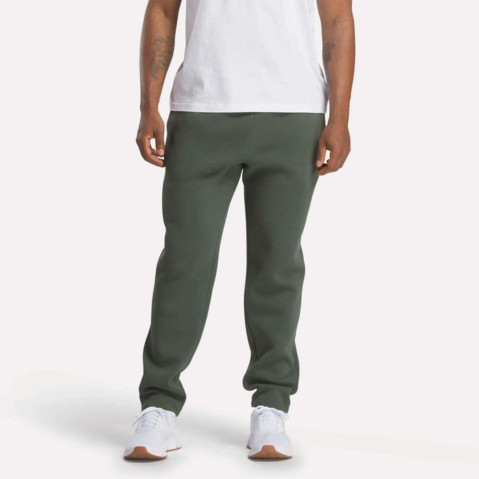 A person wearing a white t-shirt, Reebok Men's Patent Pants in slim fit olive green with an adjustable waistband, and white sneakers stands against a plain light background; their face is not visible.