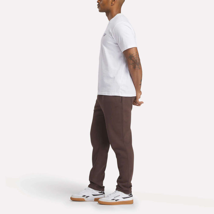 A man stands in profile against a white background, wearing a slim fit white t-shirt, Reebok Men's Patent Pants in brown, and white sneakers with brown soles. His arms are relaxed behind his back, revealing tattoos on his left forearm.