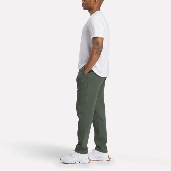A man stands in profile with hands in pockets, wearing a white t-shirt and Reebok Men's Patent Pants in slim fit olive green with an adjustable waistband, along with white sneakers, against a plain white background.