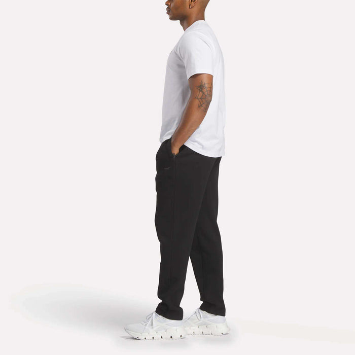 A person stands in profile against a light background, wearing a white t-shirt, Reebok Men's Patent Pants with an adjustable waistband, and white sneakers. Their left hand is in their pocket, revealing a geometric tattoo on their upper right arm.