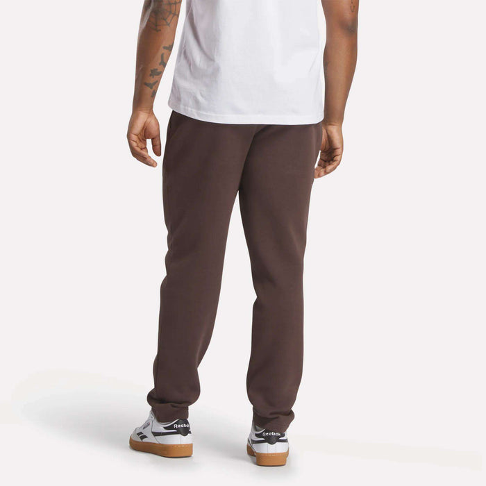 A person stands with their back to the camera, wearing a white t-shirt and slim fit dark brown Men's Patent Pants by Reebok, made from a cotton-poly blend, with white sneakers. Their tattooed right arm is visible against a plain light background.