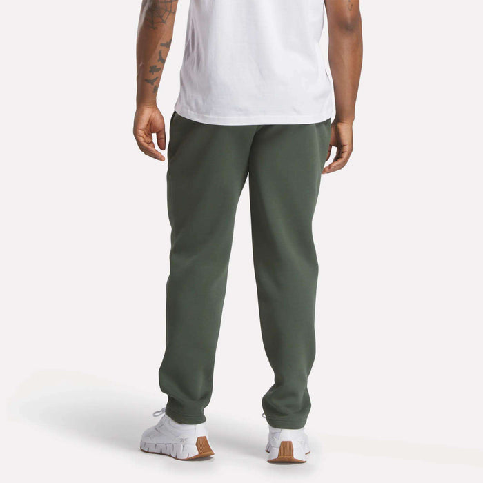 A person stands facing away, wearing Reebok Men's Patent Pants in dark green with a slim fit and adjustable waistband. Only their lower back, legs, and part of the arms are visible against a plain background.