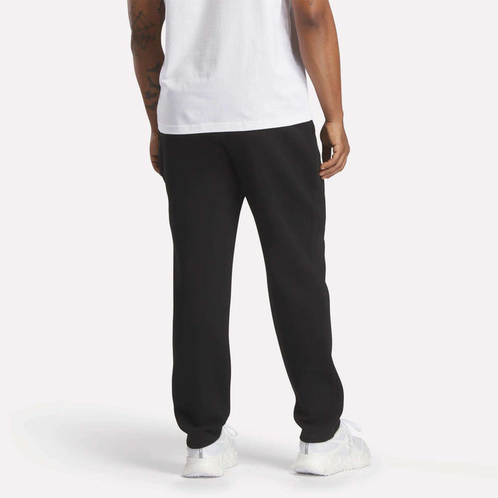 A person stands with their back to the camera against a plain white background, wearing a white t-shirt, Reebok Men's Patent Pants in black with an adjustable waistband, and white sneakers.