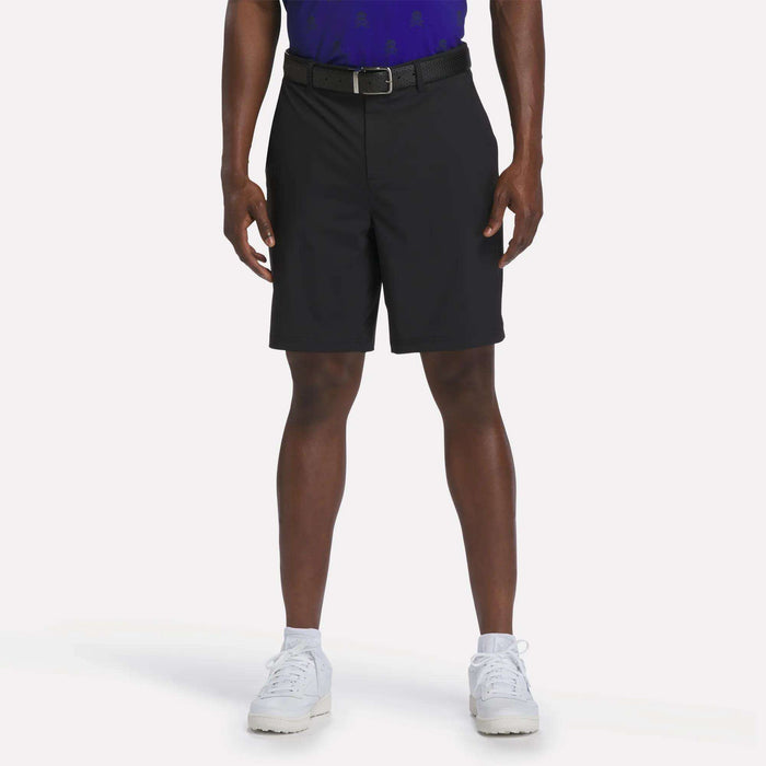 A person stands against a plain white background wearing a blue shirt and the Reebok Men's Pull-On Golf Shorts 8.5" in black, which feature four-way stretch for added comfort. The look is completed with white sneakers.