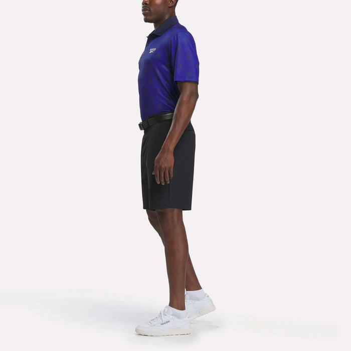 A man stands sideways against a plain white background, wearing a blue collared shirt and Reebok Men's Pull-On Golf Shorts 8.5" in black, featuring a comfortable four-way stretch pull-on design. He pairs the outfit with white socks and white sneakers.