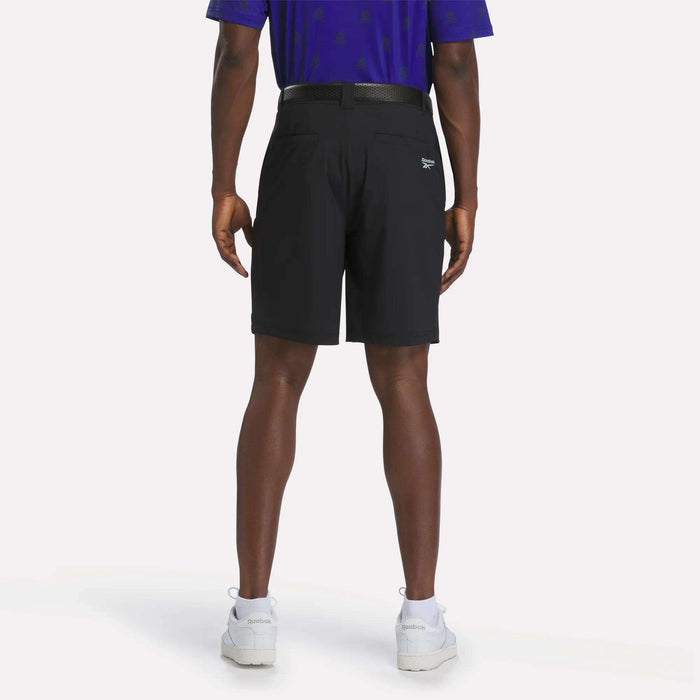 A man stands facing away, wearing a blue shirt and Reebok Men's Pull-On Golf Shorts 8.5" in black with a belt, featuring four-way stretch for comfort and ease of movement, paired with white sneakers against a plain white background.
