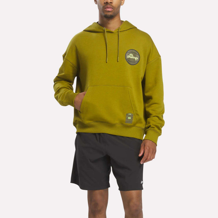 A person wearing the Reebok Men's Pump OG Hoodie in murky green, featuring a round logo on the chest, and black shorts stands with one hand in the hoodie pocket against a plain background, channeling classic retro basketball style reminiscent of the Reebok Pump era.