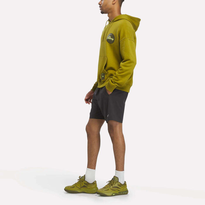 A man in a Reebok Men's Pump OG Hoodie, black shorts, white socks, and green Reebok Pump sneakers stands facing left against a plain white background.