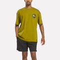 A man wearing a murky green Reebok Men's Pump OG T-Shirt featuring a round The Pump logo on the chest and black athletic shorts, standing with one hand in his pocket.