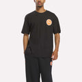 A person is wearing a loose-fitting black Reebok Men's Pump OG T-Shirt, which features an orange and white circular Pump logo, paired with black pants, standing against a plain light background.
