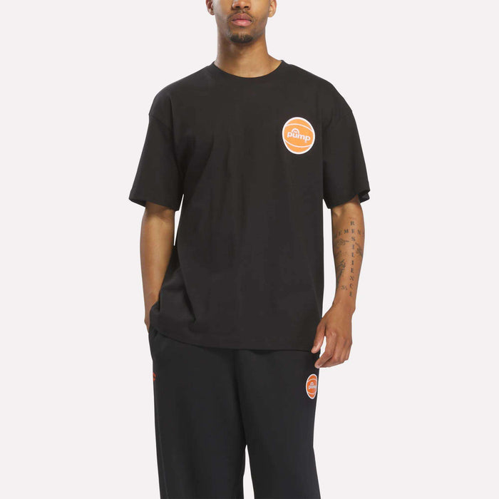 A person is wearing a loose-fitting black Reebok Men's Pump OG T-Shirt, which features an orange and white circular Pump logo, paired with black pants, standing against a plain light background.