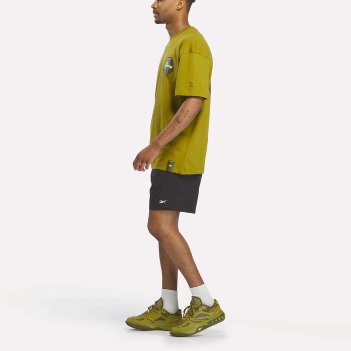 Wearing the Reebok Men's Pump OG T-Shirt in yellow-green, paired with black shorts, white socks, and green sneakers, a person stands in profile against a plain background.