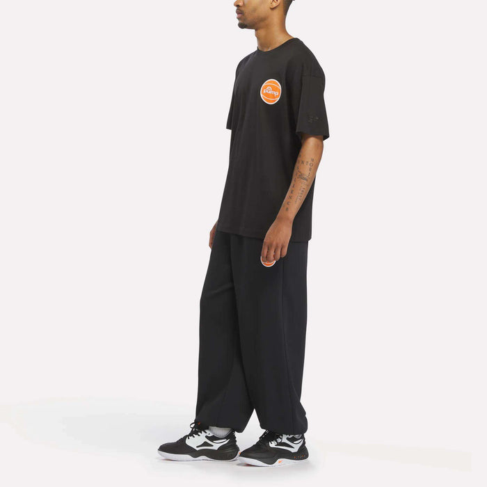 Man in a loose-fitting Reebok Men's Pump OG T-Shirt with a round orange logo, wide black pants, and Reebok Pump black-and-white sneakers standing sideways on a white background.