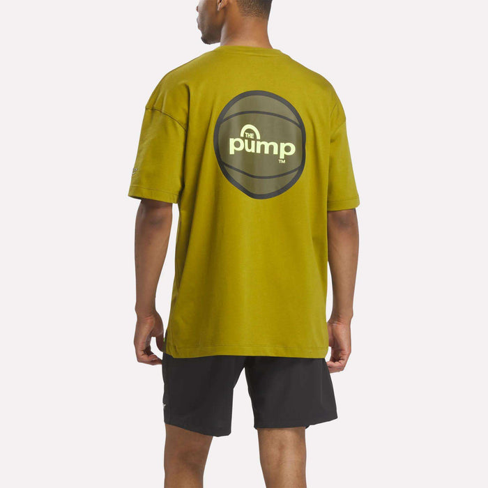 A man is wearing the Reebok Men's Pump OG T-Shirt in olive green, featuring The Pump logo on the back, paired with black shorts. He is standing facing away from the camera against a white background.