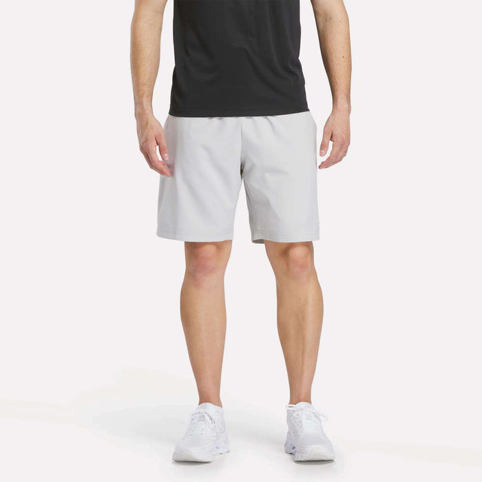 A man in a black T-shirt, Reebok Men’s Range Woven Shorts 9″ in Grey 1, and white sneakers stands facing forward against a plain white background.