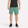 A person wearing a black t-shirt, Men’s Range Woven Shorts 9″ by Reebok in Field Green, and black sneakers stands against a plain white background. The photo is cropped at the shoulders and shows only the lower torso and legs.