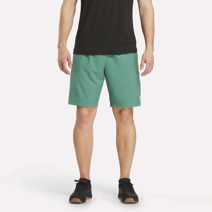 A person wearing a black t-shirt, Men’s Range Woven Shorts 9″ by Reebok in Field Green, and black sneakers stands against a plain white background. The photo is cropped at the shoulders and shows only the lower torso and legs.