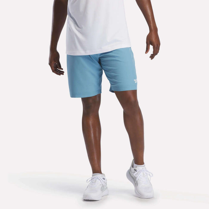 A person wearing a white shirt, Reebok Men's Range Shorts 9" in Warped Blue, and white sneakers stands against a plain white background.