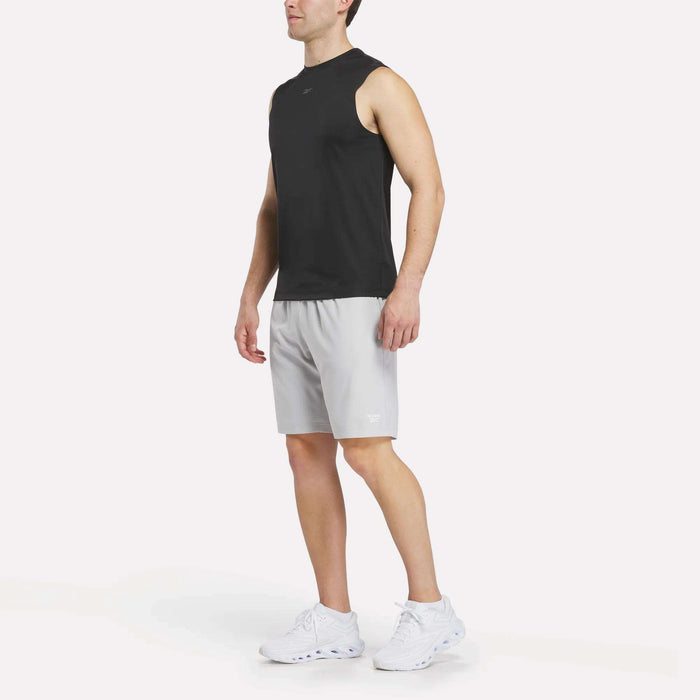 A man stands against a plain white background wearing a sleeveless black athletic shirt, white athletic shoes, and light gray Reebok Men’s Range Woven Shorts 9″.