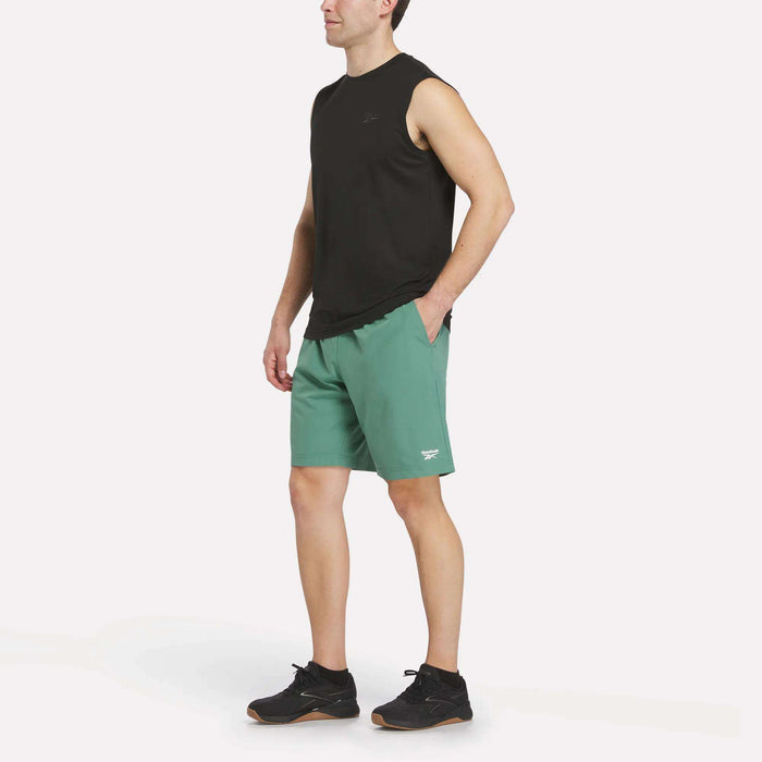 A person wearing a sleeveless black shirt, black athletic shoes, and teal green Reebok Men’s Range Woven Shorts 9″ stands against a plain white background.