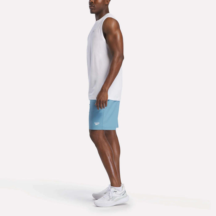A man stands sideways against a plain white background, wearing a sleeveless white shirt, white sneakers, and light blue Reebok Men's Range Shorts 9".