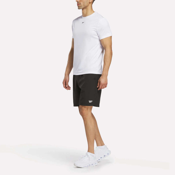 A person wearing a white short-sleeve t-shirt, Reebok Men’s Range Woven Shorts 9″ in black, and white sneakers stands against a plain white background.