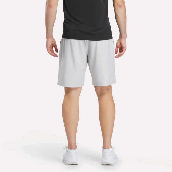 A person stands facing away, wearing a black t-shirt, white sneakers, and light gray Reebok Men’s Range Woven Shorts 9″ against a plain white background.