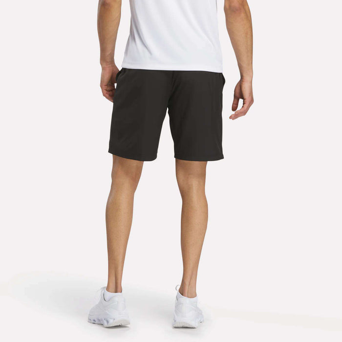 A person stands facing away, wearing a white shirt, Reebok Men’s Range Woven Shorts 9″, and white sneakers. Only the lower half of the body is visible, shown against a plain white background.